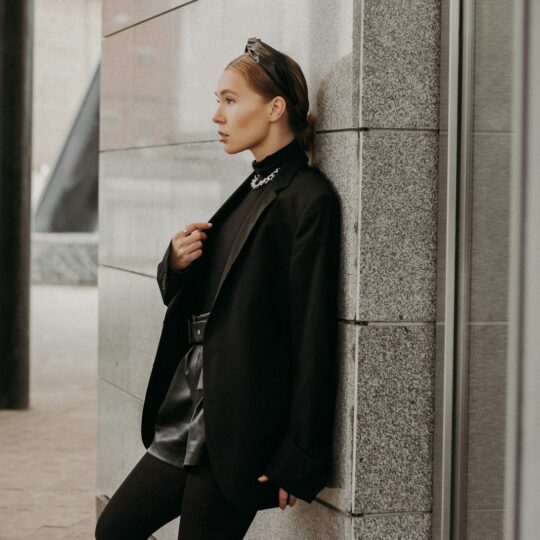 woman in black coat and black pants leaning on gray concrete wall